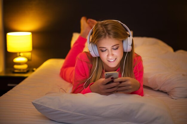 Jovens mulheres loiras sorrindo ouvindo música com fone de ouvido de um smartphone deitado na cama em casa