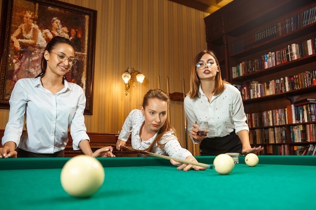 Jovens mulheres sorridentes jogando bilhar no escritório ou em casa depois do trabalho. Colegas de trabalho envolvidos em atividades recreativas. Amizade, atividade de lazer, conceito de jogo.