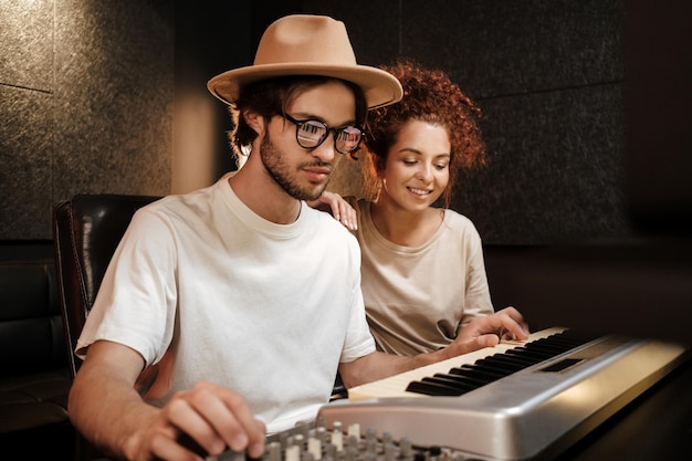 Jovens músicos elegantes gravando alegremente uma nova música no teclado de piano no estúdio de som moderno. homem criativo atraente e mulher trabalhando em novo álbum de música