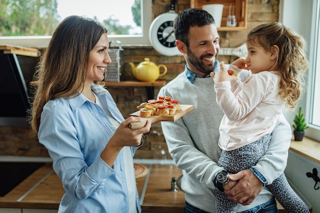 Jovens pais felizes e sua filha pequena se comunicando enquanto comem bruschetta na cozinha