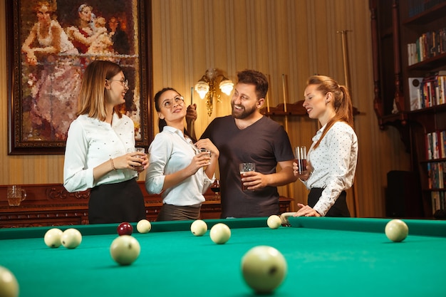 Jovens sorrindo, homens e mulheres jogando bilhar no escritório ou em casa, depois do trabalho. Colegas de trabalho envolvidos em atividades recreativas.