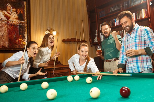 Jovens sorrindo, homens e mulheres jogando bilhar no escritório ou em casa, depois do trabalho.