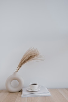 Junco fofo de capim-dos-pampas em um vaso estiloso, xícara de café e revistas contra a parede branca