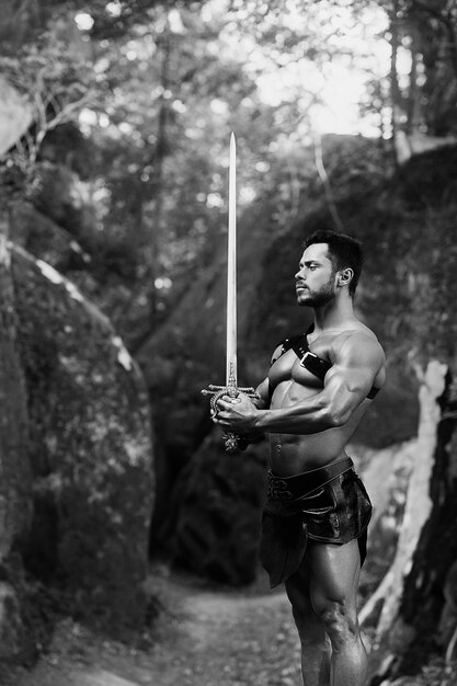 Justiça é sua única regra. Foto monocromática vertical de um jovem gladiador forte e corajoso segurando uma espada em pé perto das rochas na floresta