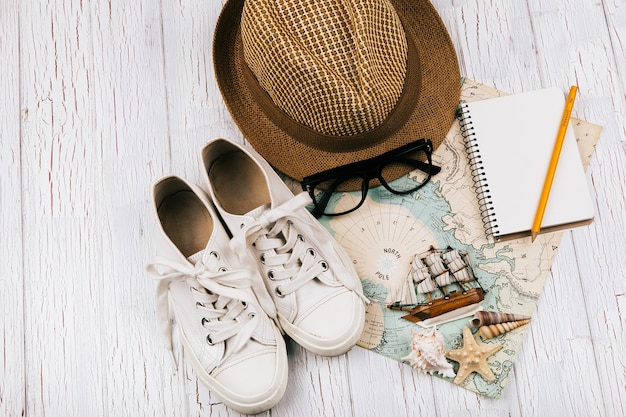 Keds, chapéu, óculos, caderno, pequeno navio de madeira se encontram no branco mapa de viagens