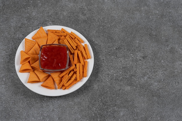 Ketchup, salgadinhos de milho e pão seco no prato na superfície do mármore