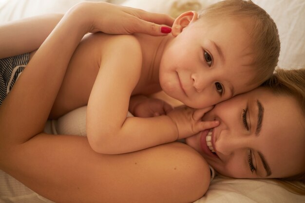 Laços familiares e conceito de maternidade. Imagem recortada da bela jovem mãe europeia relaxando no quarto, deitada em lençóis brancos com seu adorável filho bebê, segurando-o com força e sorrindo alegremente