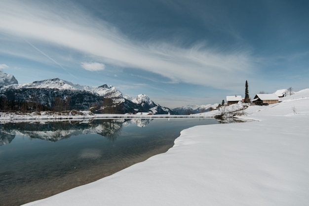 Lago cercado por pedras e casas cobertas de neve sob o sol