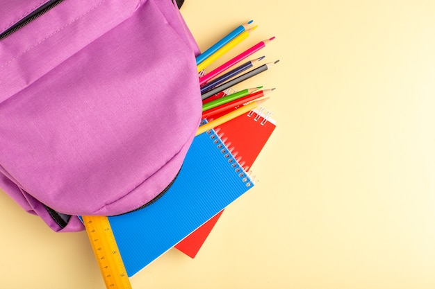 Lápis coloridos de vista superior com cadernos e bolsa roxa no bloco de notas de caneta escolar de parede amarelo-claro.