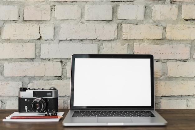 Laptop com tela branca em branco perto da câmera; bloco de anotações; lápis e buldogue na mesa de madeira