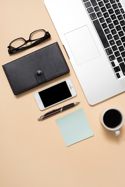 Laptop de imagem recortada com óculos; celular; xícara de café e diário sobre fundo bege