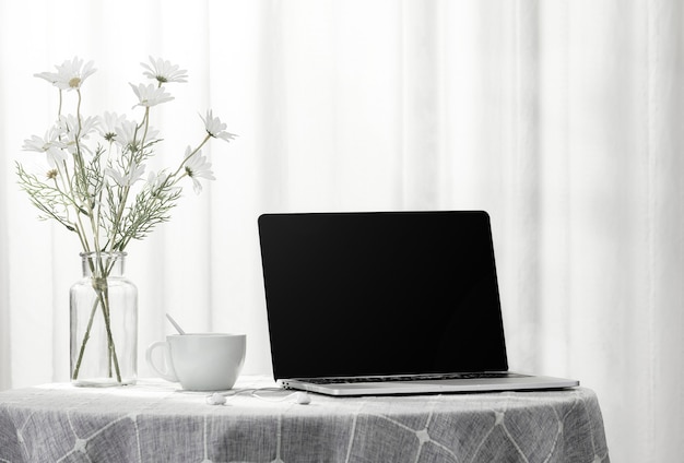 Laptop e uma xícara de café ao lado de um vaso cheio de flores, dentro de casa