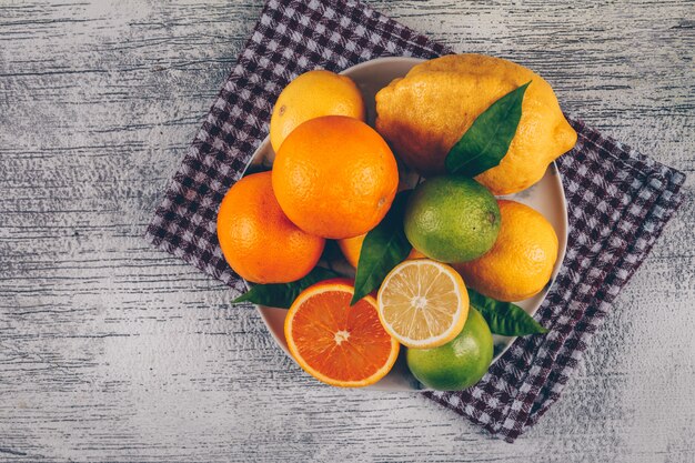 Laranja com limões verdes e amarelos com fatias em uma placa no pano e no fundo de madeira cinzento, vista superior.