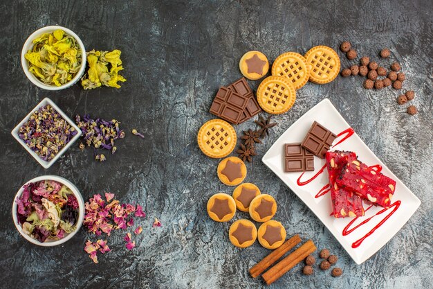 Layout em forma de crescente de doces com um prato de chocolates e tigelas de flores secas em um fundo cinza