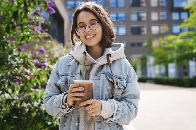 Lazer de estilo de vida e conceito de atividade ao ar livre Retrato de menina queer feliz na jaqueta jeans e óculos dando um passeio com a cidade aproveitando a primavera bebendo café com leite e câmera sorridente