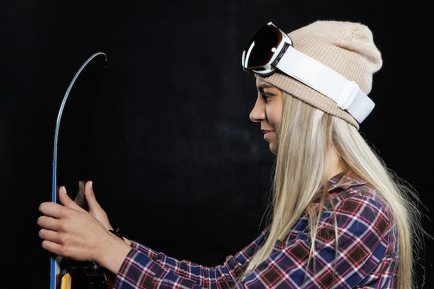 Lazer de inverno, esportes radicais e conceito de atividade. Retrato do perfil do snowboarder jovem loira feliz usando chapéu e máscara de esqui, posando dentro de casa com snowboard preto, vestindo-se para andar