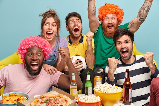 Lazer, esporte, conceito de felicidade. Amigos emocionados e felizes levantam as mãos, gritam bem alto, comemoram o gol, estão contentes com a vitória do popular time de futebol, fazem um lanche, bebem bebida alcoólica, posam dentro de casa