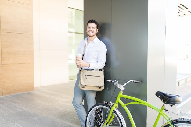 Legal jovem em trajes formais com xícara de café e bicicleta sorrindo no corredor