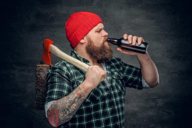 Lenhador barbudo brutal vestindo camisa xadrez verde e chapéu vermelho, bebendo cerveja de uma garrafa e segura o machado no ombro.