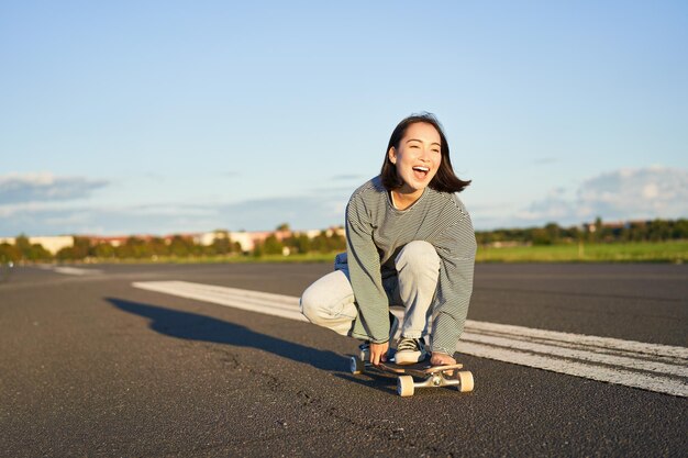 Liberdade e felicidade menina asiática feliz montando seu longboard em uma estrada ensolarada vazia rindo e sorrindo