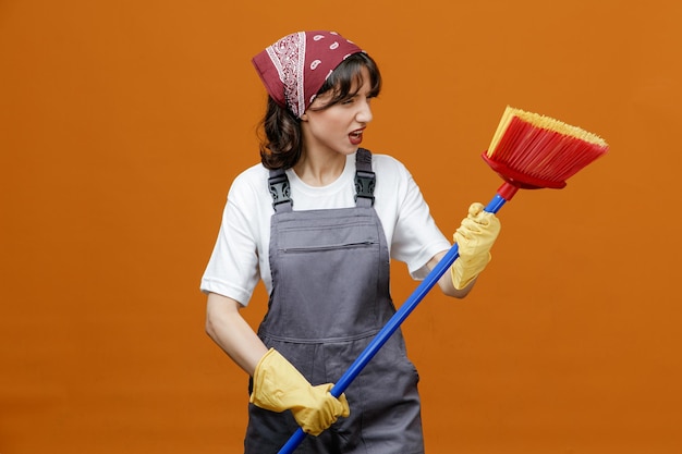 Limpador feminino jovem irritado usando luvas de borracha uniforme e bandana segurando o esfregão de rodo com as duas mãos olhando para o lado isolado em fundo laranja