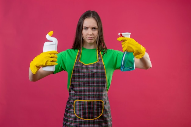 Limpando jovem mulher usando uniforme com luvas segurando agente de limpeza e spray na parede rosa isolada