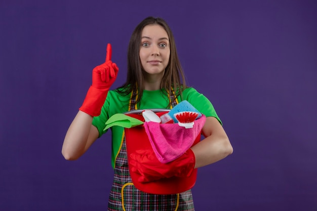 limpando mulher jovem usando uniforme com luvas vermelhas segurando ferramentas de limpeza apontando o dedo para cima na parede roxa isolada