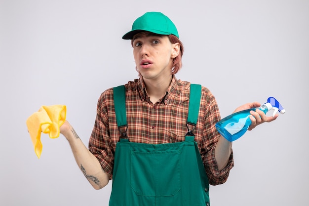 Limpeza jovem com macacão e boné de camisa xadrez, segurando um pano e spray de limpeza, parecendo surpreso e confuso