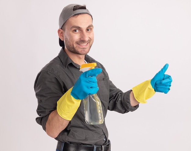 Foto grátis limpeza jovem vestindo roupas casuais e boné em luvas de borracha segurando spray de limpeza e sorrindo e mostrando os polegares em pé sobre a parede laranja