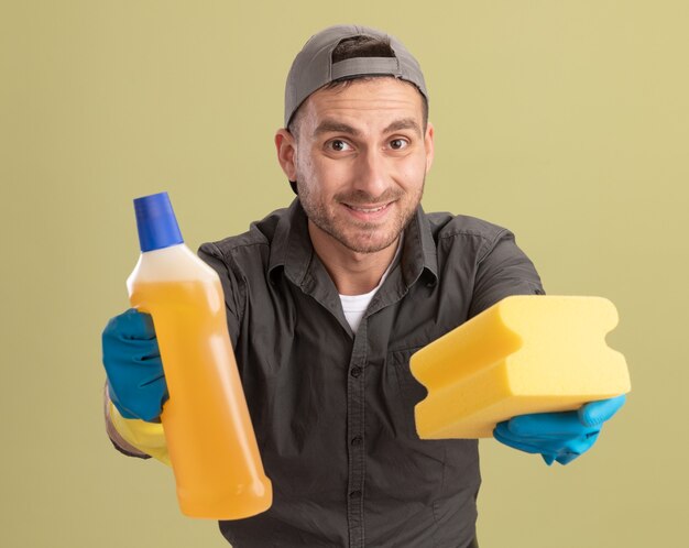 Limpeza jovem vestindo roupas casuais e boné em luvas de borracha, segurando uma garrafa com material de limpeza e uma esponja, olhando sorrindo em pé sobre a parede verde