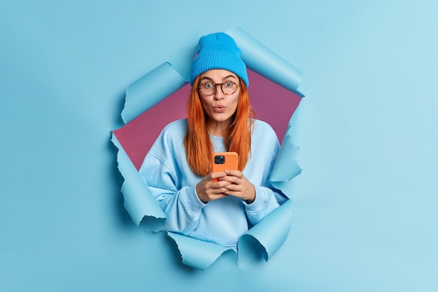 Foto grátis linda adolescente ruiva lê postagem em redes sociais, bate-papos online, usa celular prende a respiração parece maravilhada usa gadget moderno vestida com roupas elegantes quebra a parede de papel