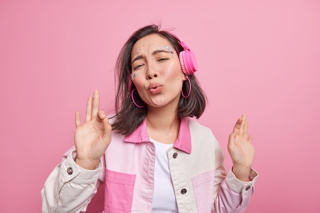 Linda alegre adolescente asiática dança em fones de ouvido sem fio modernos, mantém os lábios dobrados, desfruta da melodia de cantar de música favorita mantém as mãos levantadas isoladas sobre a parede rosa. O amante da música se diverte em casa