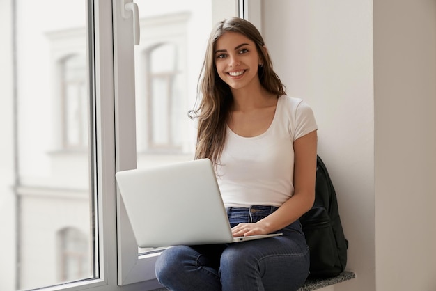 Linda aluna sentada com laptop no peitoril da janela