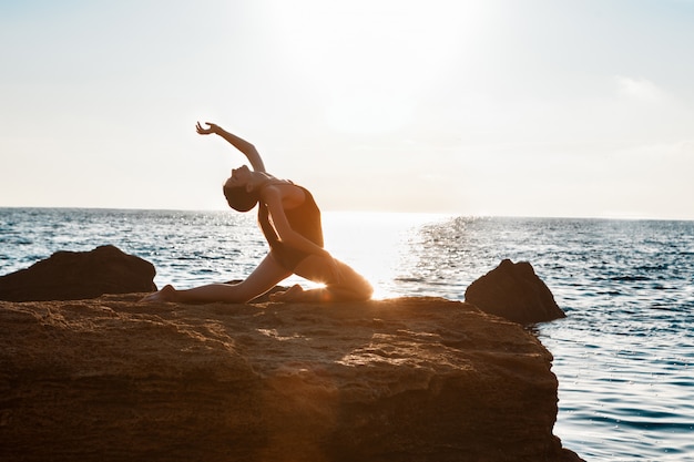 Linda bailarina dançando, posando em pedra na praia, vista para o mar.