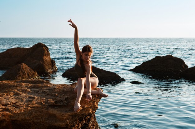 Linda bailarina dançando, posando em pedra na praia, vista para o mar.