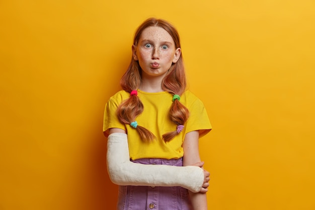 Linda colegial faz uma expressão engraçada, faz beicinho, tem dois rabos de cavalo, cabelo ruivo, rosto sardento, vestida casualmente, se machucou após cair de altura, usa gesso no braço quebrado, isolado no amarelo