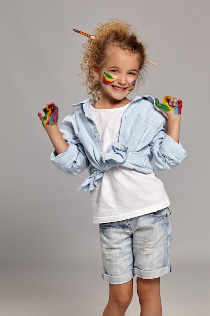 Linda criança com uma escova em seu penteado elegante, vestindo uma camisa azul e camiseta branca. ela está posando com os dedos pintados e sorrindo amplamente para a câmera, em um fundo cinza.