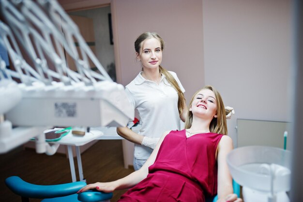 Linda dentista feminina posando e sorrindo com sua adorável paciente deitada em uma cadeira odontológica com vestido vermelho-violeta