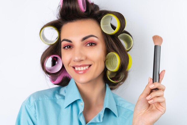 Linda dona de casa Jovem alegre com maquiagem rosa brilhante de rolos de cabelo e uma escova de contorno está se preparando para um jantar noturno em um fundo branco