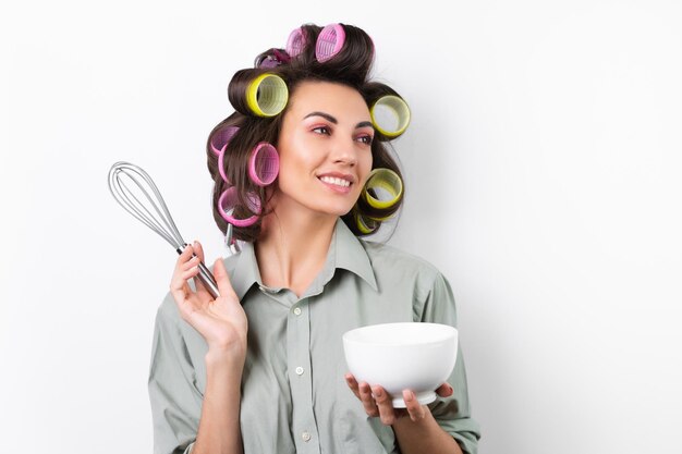 Linda dona de casa jovem alegre com rolinhos de cabelo maquiagem brilhante uma xícara branca e um batedor nas mãos em um fundo branco Pensando na receita para o jantar