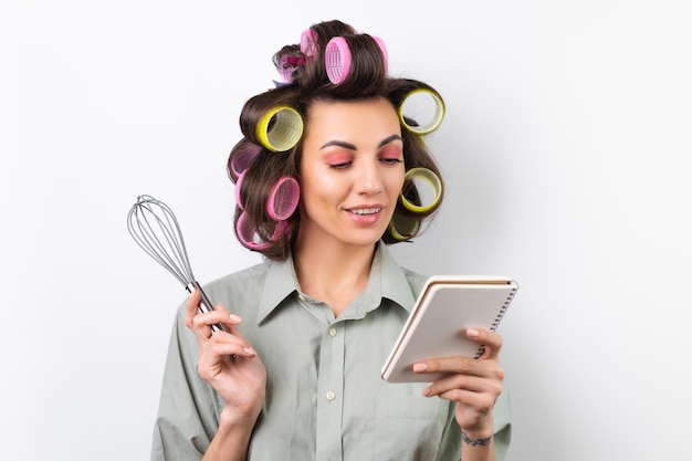 Linda dona de casa jovem alegre com rolos de cabelo maquiagem brilhante um caderno e um batedor nas mãos em um fundo branco Pensando na receita para o jantar