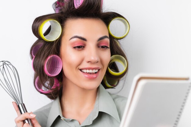 Linda dona de casa jovem alegre com rolos de cabelo maquiagem brilhante um caderno e um batedor nas mãos em um fundo branco Pensando na receita para o jantar