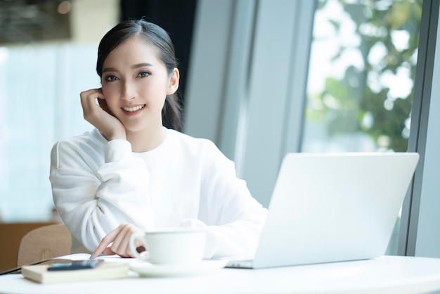 Linda e atraente frescura feminina asiática sorria alegre manhã trabalhando com café portátil trabalho em casa com felicidade aproveite a vista para o jardim jovem asiática com janela luz de fundo do jardim
