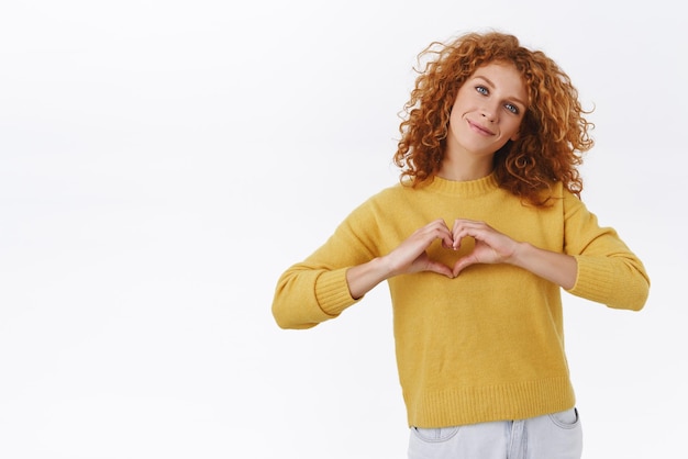 Linda e encantadora mulher encaracolada ruiva de suéter amarelo absolutamente apaixonada pelo café favorito expressa amor ao café fazer sinal de coração sobre o peito inclinar a cabeça e sorrir mostrar carinho e ternura