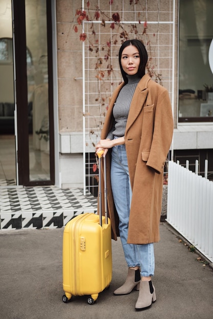Linda e estilosa garota asiática de casaco sonhadoramente olhando para longe em pé com mala amarela na rua da cidade