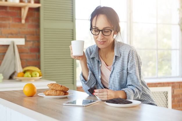 Linda e jovem modelo feminina usa óculos e camisa, toma café com croissants e chocolate amargo, toma café da manhã antes do trabalho,