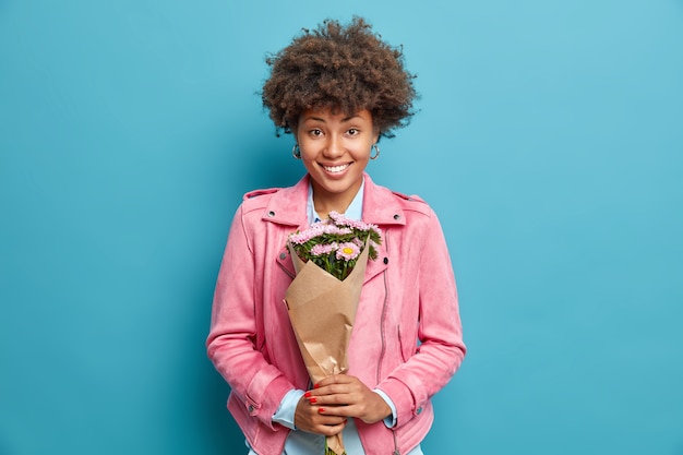 Linda e linda mulher afro-americana com cabelo encaracolado segurando um buquê de flores indo parabenizar a melhor amiga no feriado, tem um clima festivo e feliz, usa uma jaqueta rosa isolada na parede azul do estúdio