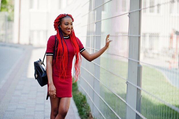 Linda e magra garota afro-americana de vestido vermelho com dreadlocks e mochila posada ao ar livre em portões de gaiola de fundo de rua Modelo preto elegante