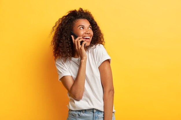 Linda e satisfeita jovem afro-americana gosta de uma conversa agradável, segura o celular perto da orelha, olha para o lado e usa uma camiseta branca casual