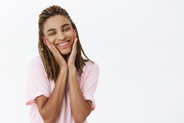 Linda e terna garota elegante de pele escura com dreads e aparelho, sorrindo alegremente segurando as palmas das mãos nas bochechas, sentindo-se bonita e encantada